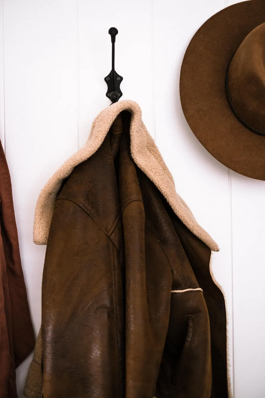 A brown shearling-collared leather jacket and a felt hat hanging on wall hooks.