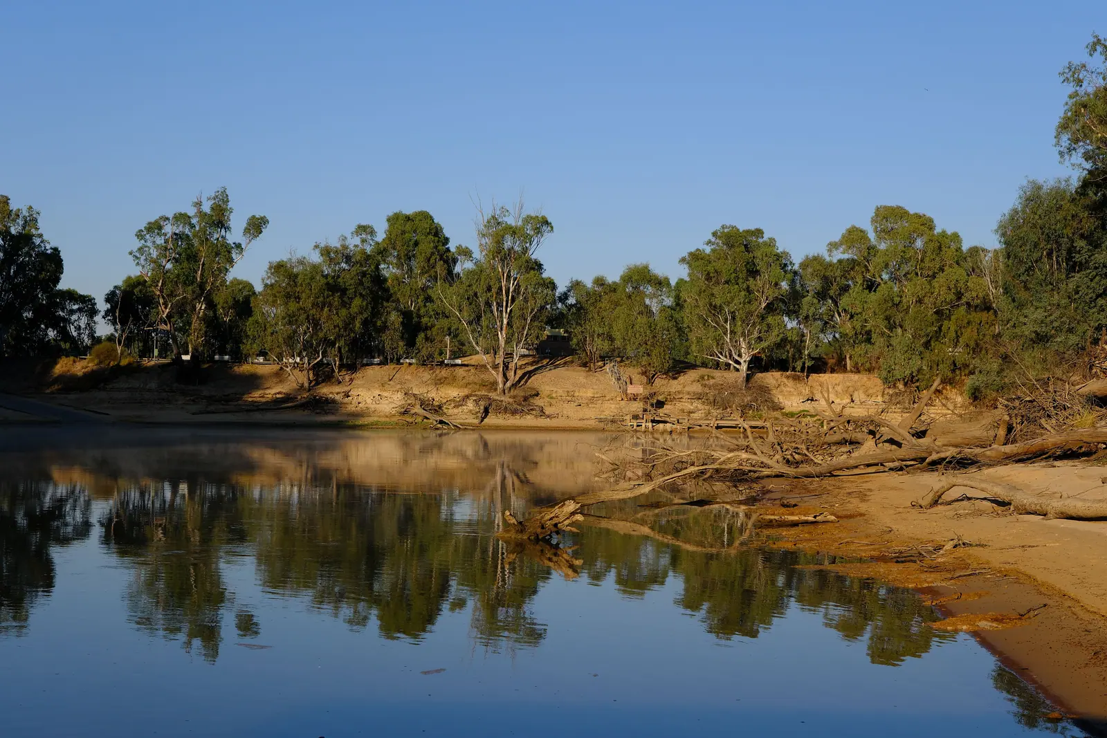 A still inland river reflecting gum trees and sandy banks under a clear sky.